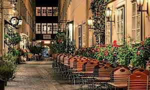 En pittoresk gate med stoler og bord utenfor restauranter, omgitt av blomsterbusker og lamper. (AI-generert tekst)