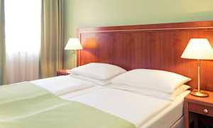 Et hotellrom med en dobbeltseng som har hvite sengeklær og grønne tepper. To sengelamper står på trebord ved sengen. (AI-generert tekst)