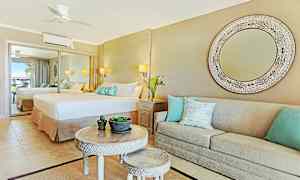 Et elegant hotellrom med et stort speil, en bred seng og en beige sofa med dekorative puter. Et rundt bord med mindre bord og plantepynt i midten. (AI-generert tekst)