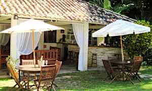 Utendørsmiljø med bord og stoler av tre samt parasoller, ved siden av en bygning med takstein og innvendige gardiner. (AI-generert tekst)