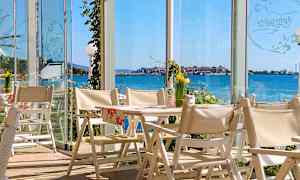 Utsikt fra en restaurant med hvite stoler og bord, med glassvegger som gir utsyn over havet og en kystby. (AI-generert tekst)