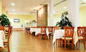 Bilde av en spisesal med bord og stoler i tre, hvite duker, planter og en bufféseksjon. (AI-generert tekst)