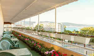 En terrasse med grønne plaststoler og bord, omgitt av blomsterpotter og utsikt mot et hotell og havet. (AI-generert tekst)