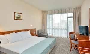 Et hotellrom med en dobbeltseng, skrivebord, stol, TV og sittegruppe. Et stort vindu slipper inn dagslys. (AI-generert tekst)