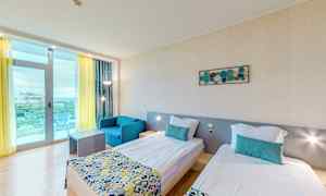 Et hotellrom med to enkeltsenger, en blå lenestol og et bord. Rommet har lyse vegger og et stort vindu med gul gardin og balkong. (AI-generert tekst)