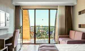 Et lyst hotellrom med balkong, møblert med seng, sofa og skrivebord. (AI-generert tekst)