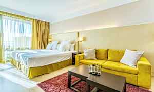 Et hotellrom med en stor dobbeltseng, gul sofa, et bord med dekorativ vase og et rødt teppe. (AI-generert tekst)