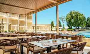 Utendørsrestaurant med bord og stoler under tak, omgitt av grønt og et basseng i bakgrunnen. (AI-generert tekst)