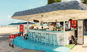 En strandbar med blå stoler og tak, plassert nær sandstranden. (AI-generert tekst)