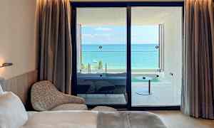 Et hotellrom med en seng og lenestol, samt utsikt over en balkong med havet i bakgrunnen. (AI-generert tekst)