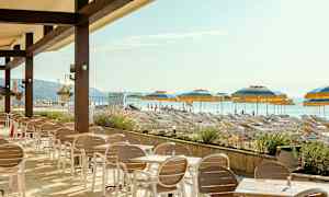 En uteservering med bord og stoler foran en strand med solsenger og parasoller. (AI-generert tekst)