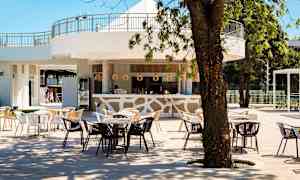 En utendørsbar med bord og stoler under trær, med lys innredning og tre detaljer. (AI-generert tekst)