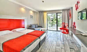 Et hotellrom med to enkeltsenger, en sofa, en skrivebordplass og en balkong med utsikt mot grønne omgivelser. (AI-generert tekst)