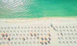 Flyfoto av en strand med parasoller og solsenger. Havet er tiltalende grønnblått. (AI-generert tekst)
