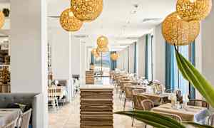 En moderne restaurant med en lys innredning som inkluderer trebord, stoler og store vinduer med blå gardiner. Dekorative belysningslamper henger fra taket. (AI-generert tekst)