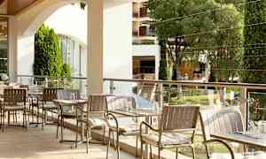 En utendørs serveringsområde med bord og stoler, omgitt av grønne trær og beplantning. (AI-generert tekst)