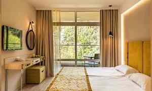 Et hotellrom med en dobbeltseng, gul sengegavl, lite skrivebord, og en stor vindusfront mot naturen. (AI-generert tekst)