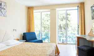 Rom med seng, blå lenestol og skrivebord, med store vinduer mot balkong. (AI-generert tekst)