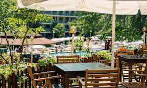 En uteplass med bord og stoler av tre under en parasoll, ved en hage og bassengområde. (AI-generert tekst)