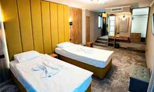 Et hotellrom med to enkeltsenger med gul polstring og hvitt sengetøy. Rommet har en moderne innredning med en gul veggpanel, et skrivebord, sittemøbler og en liten oppbevaringshylle. (AI-generert tekst)