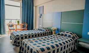 To enkeltsenger i et rom med blå og grå innredning. Et lite bord med to stoler står ved et stort vindu. (AI-generert tekst)