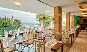 En restaurant med bord og stoler i lyse farger, utsikt over havet og omgitt av trær. (AI-generert tekst)