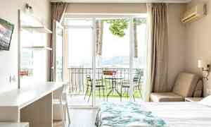 Et hotellrom med en seng, et bord og en stol, samt en balkong med utemøbler og utsikt mot naturen. (AI-generert tekst)