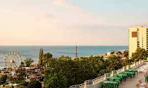 Bildet viser en kystlinje med havet, et pariserhjul, frodige trær og bygninger. (AI-generert tekst)