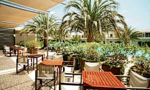 En uteservering med bord og stoler nær et basseng omgitt av palmer. (AI-generert tekst)