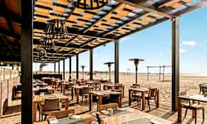 Utendørsrestaurant med trebord og stoler, plassert ved en sandstrand. (AI-generert tekst)