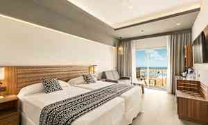 Et hotellrom med to senger og en sittegruppe ved siden av en glassdør som fører til en balkong med utsikt. (AI-generert tekst)