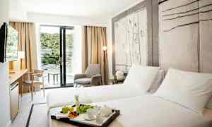 Et hotellrom med to enkeltsenger, en sittegruppe og en balkong med utsikt over et frodig område. (AI-generert tekst)