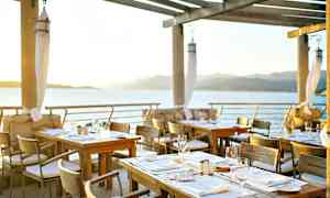 Uteservering med oppstilte bord ved vannet, med utsikt over havet og fjellene i solnedgangen. (AI-generert tekst)