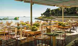 Uteservering ved sjøen med bord og stoler under et tak, omgitt av planter og en strand i bakgrunnen. (AI-generert tekst)