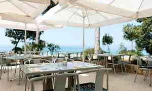 En uteservering med hvite parasoller, bord og stoler samt utsikt over havet. (AI-generert tekst)