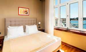 Et hotellrom med en dobbeltseng, hvite puter, beige sengeteppe og et stort vindu med utsikt mot havet. (AI-generert tekst)