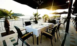 Uteservering med bord og stoler, med havutsikt og solnedgang. (AI-generert tekst)