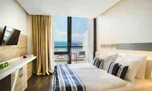 Et hotellrom med en dobbeltseng, sengetøy i blått og hvitt stripemønster, og en stor glassvegg som viser en balkong med utsikt over havet. Rommet har også en TV og skrivebord. (AI-generert tekst)