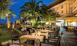 Kveldsbilde av en utendørsrestaurant med opplyste bord, omgitt av palmer og grøntområder. (AI-generert tekst)