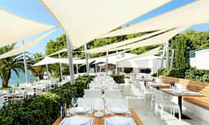 Et utendørsområde med flere bord og stoler under spennende parasoller nær vannet. (AI-generert tekst)