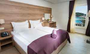 Et hotellrom med en dobbeltseng, hvite laken og lilla sengeteppe. Det er et skrivebord med en stol og et speil. Vinduet viser utsikt mot havet. (AI-generert tekst)