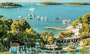 Utsikt over et bassenganlegg med solstoler omgitt av grønn vegetasjon. I bakgrunnen sees en strand med båter og en bukt med små øyer. (AI-generert tekst)