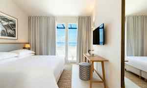 Et hotellrom med en stor seng, et skrivebord og en TV på veggen. Store vinduer med utsikt mot havet. (AI-generert tekst)