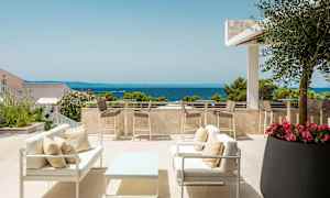 En terrasse med sittemøbler og bord samt utsikt over havet. Den er omgitt av planter og trær med klar himmel. (AI-generert tekst)