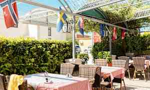 Utendørsterrasse med bord, stoler og flagg, omgitt av grønne planter. (AI-generert tekst)