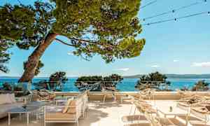 Utendørs terrasse med stoler og bord, med utsikt mot havet og omkringliggende trær. (AI-generert tekst)