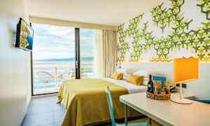 Hotellrom med moderne innredning, gul sengeteppe, grønn mønstret vegg og utsikt over havet fra balkongen. (AI-generert tekst)