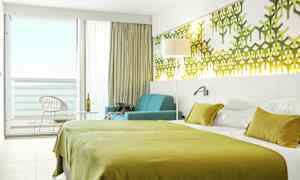 Et hotellrom med grøn sengetrekk, veggdekorasjoner og en sittegruppe ved et vindu mot balkongen. (AI-generert tekst)