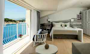 Et hotellrom med en oppredd seng, sitteområde og en balkong med utsikt over havet. (AI-generert tekst)