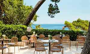 Bilde av en uteservering med bord og stoler, omgitt av grøntområder og med havutsikt. (AI-generert tekst)